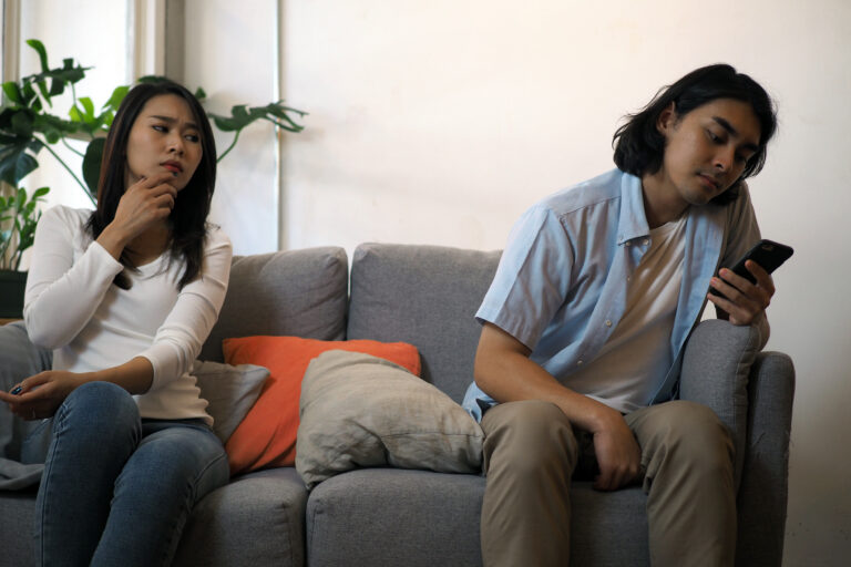 Two people sit on a gray sofa indoors; the woman on the left looks pensive while touching her chin, and the man on the right glances at a smartphone in his hand.