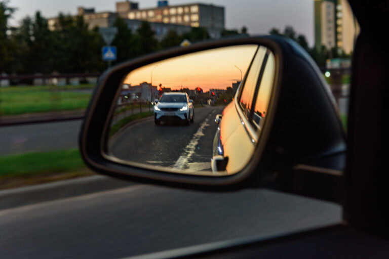 Reflection in a car's side mirror shows a sunset-lit street with an oncoming white SUV and urban background.