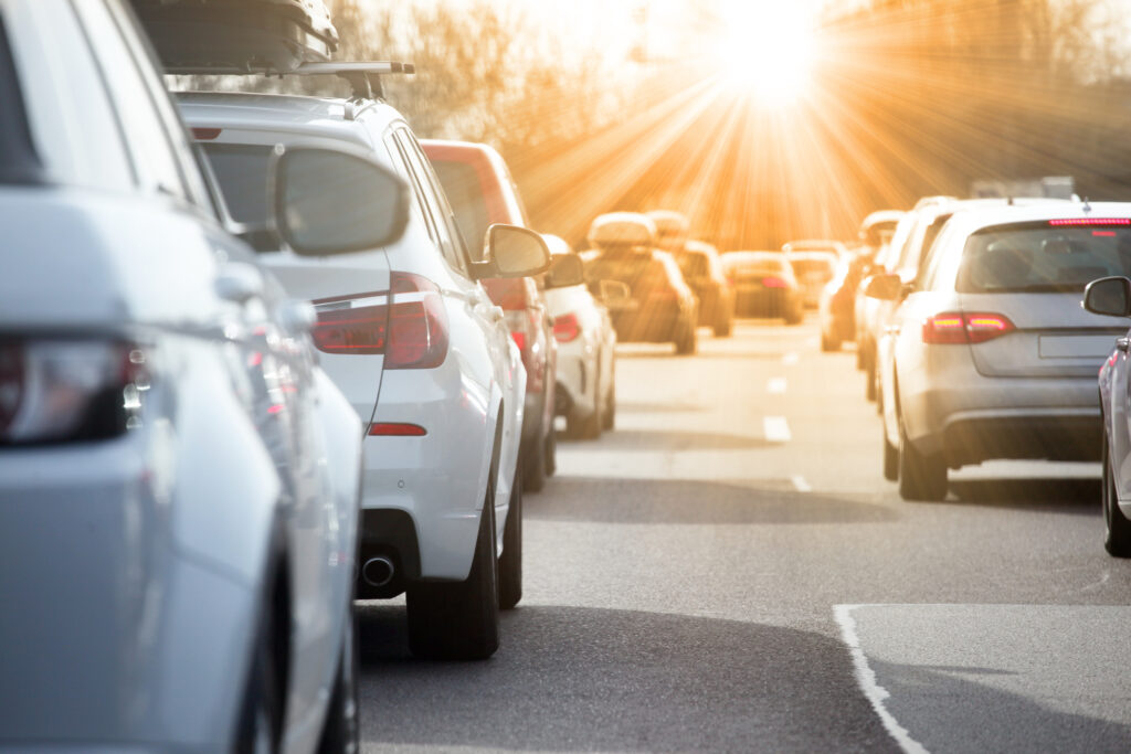 Line of cars stuck in heavy traffic on a sunlit highway, with a bright sun flare in the distance and long shadows on the road.